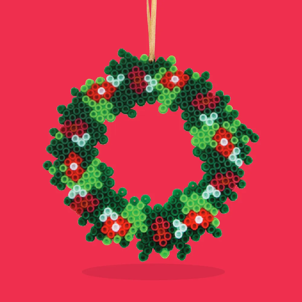 A handmade circular Christmas wreath made from green, red, and white fuse beads arranged in a holly pattern and strung with a golden ribbon for hanging.
