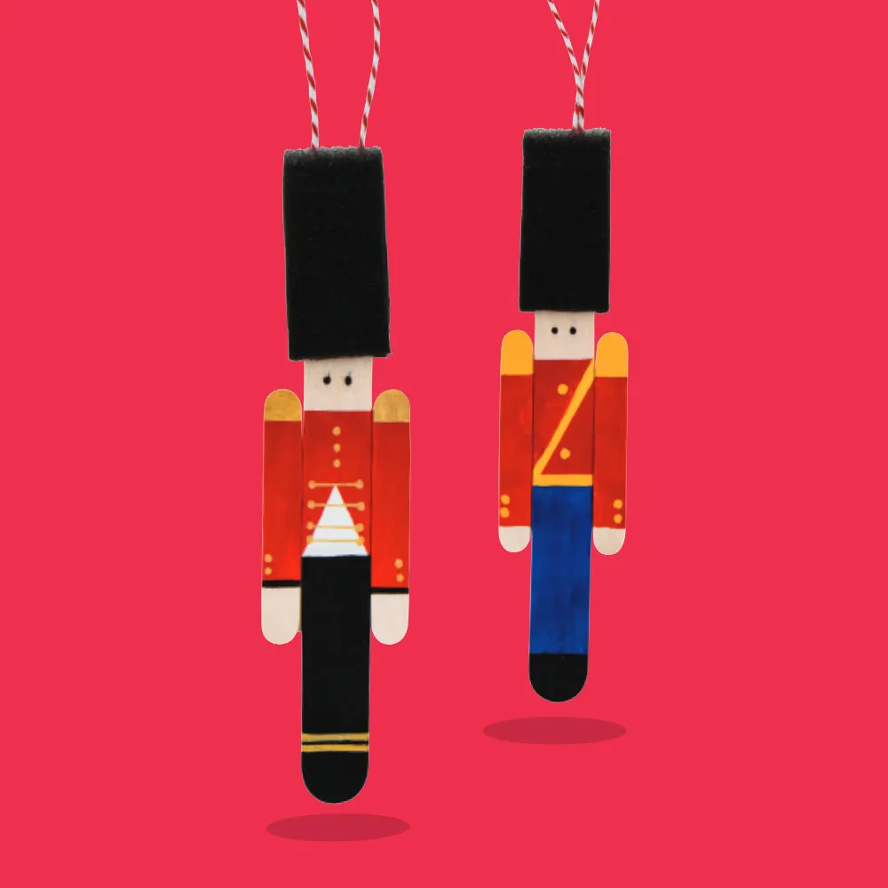 Two handcrafted nutcracker soldier ornaments made from wooden craft sticks, painted in traditional red and black uniforms with tall black hats and string for hanging.
